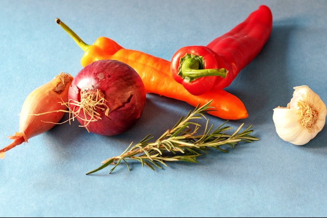 An arrangement of fresh vegetables and herbs on a blue background, including a red onion, garlic bulb, red and orange peppers, and a sprig of rosemary.