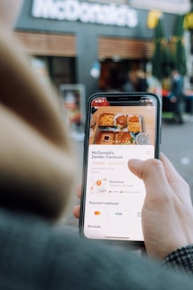 A person holds a smartphone displaying a fast-food meal order with items like fries and a burger visible on the screen. The background shows the facade of a McDonald's restaurant.