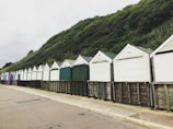A row of beach huts with peaked roofs stands along a paved path, set against a backdrop of lush, green hillside. The huts are painted in varying muted pastel shades and have closed fronts, suggesting they are currently unoccupied.