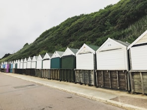 A row of beach huts with peaked roofs stands along a paved path, set against a backdrop of lush, green hillside. The huts are painted in varying muted pastel shades and have closed fronts, suggesting they are currently unoccupied.