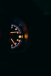A close-up view of a car dashboard displaying two circular gauges. The fuel gauge on the left is near empty, and there's a red warning light on. The other gauge is for engine temperature. The background is mostly dark, emphasizing the illuminated dials.