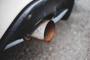 A close-up view of a car's exhaust pipe, showing a rusty, metallic texture against the smooth, dark surface of the vehicle's bumper. The image's focus is on the contrast between the weathered pipe and the polished car body.