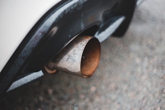 A close-up view of a car's exhaust pipe, showing a rusty, metallic texture against the smooth, dark surface of the vehicle's bumper. The image's focus is on the contrast between the weathered pipe and the polished car body.