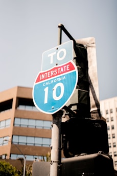 A road sign indicating directions to Interstate 10 in California, positioned on a metal pole. The sign is blue with white text and includes a red section labeled 'INTERSTATE'. In the background, there are buildings with a modern architectural style, featuring large glass windows.