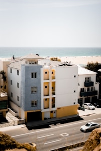 A modern multi-story building stands along a coastal road, featuring white and light blue exteriors with yellow accents. A car drives on a road in the foreground while another is parked nearby. In the background, a sandy beach and the calm blue ocean stretch into the distance under a clear sky.