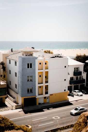 A modern multi-story building stands along a coastal road, featuring white and light blue exteriors with yellow accents. A car drives on a road in the foreground while another is parked nearby. In the background, a sandy beach and the calm blue ocean stretch into the distance under a clear sky.