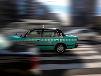 A green taxi is captured in motion against a blurred urban background, suggesting speed and dynamic city life. The taxi has a checkered stripe and a visible roof light, indicating it's a public service vehicle.