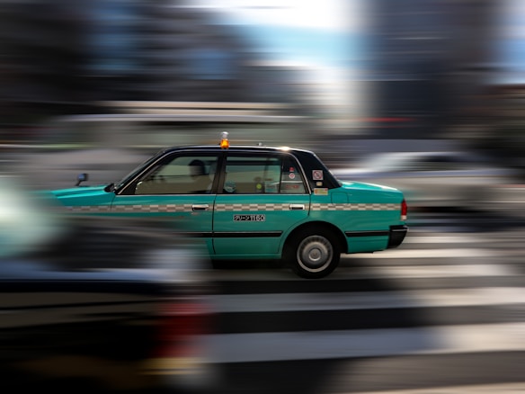 A green taxi is captured in motion against a blurred urban background, suggesting speed and dynamic city life. The taxi has a checkered stripe and a visible roof light, indicating it's a public service vehicle.