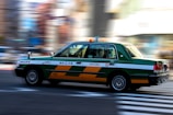 A green and white taxi is in motion, captured with motion blur effect on the background. The taxi is mid-drive on a city street, emphasizing speed and movement.