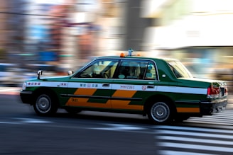 A green and white taxi is in motion, captured with motion blur effect on the background. The taxi is mid-drive on a city street, emphasizing speed and movement.