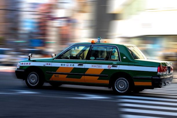 A green and white taxi is in motion, captured with motion blur effect on the background. The taxi is mid-drive on a city street, emphasizing speed and movement.