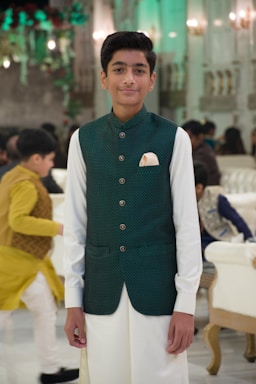 A young person is standing in a well-decorated indoor setting wearing a traditional outfit consisting of a green vest and white attire. The background features ornate decorations, elegant lighting, and other people dressed in traditional clothing, suggesting a formal or celebratory event.