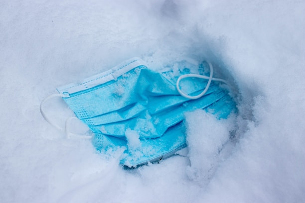 A blue disposable face mask partially covered with snow, creating an impression of abandonment or carelessness. The mask lies crumpled amidst the icy surroundings.