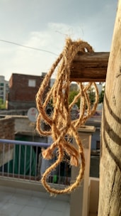 A piece of twisted jute rope is hanging on a wooden pole with a blurred urban background including rooftops, a satellite dish, and a railing. The scene is captured in bright daylight, highlighting the texture of the rope and casting shadows.