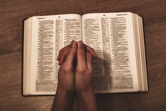 An open book, likely a Bible, placed on a wooden table with two hands clasped together in a prayer position resting over the open pages.