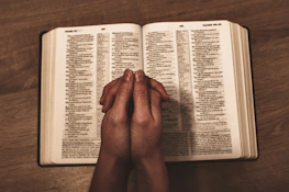 Hands joined together in prayer over an open Bible on a wooden table.