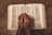 An open book, likely a Bible, placed on a wooden table with two hands clasped together in a prayer position resting over the open pages.