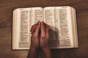 An open book, likely a Bible, placed on a wooden table with two hands clasped together in a prayer position resting over the open pages.