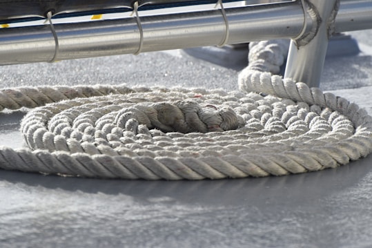 A thick white rope is coiled neatly on a ship's deck. The rope is textured and appears heavy-duty, designed for maritime use. Above the coil, there is a metal railing or support structure partially visible.