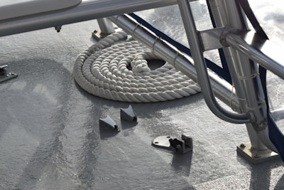 A neatly coiled white rope lies on a reflective, shiny surface near a series of metal supports and fittings, suggesting a nautical context. The metallic elements and clean, organized appearance suggest a functional and ready-to-use setting.