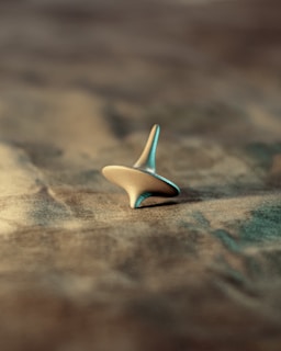 A metallic spinning top is balanced on a textured surface, with a soft focus in the background. The lighting casts a gentle shadow, highlighting the smooth and reflective edges of the top.