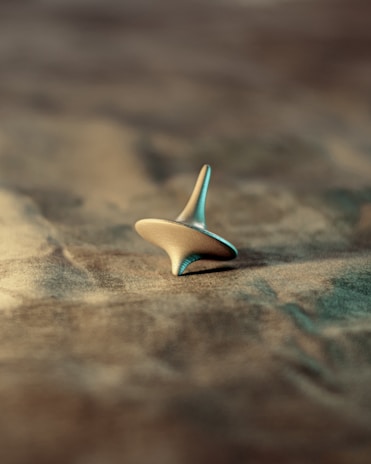 A metallic spinning top is balanced on a textured surface, with a soft focus in the background. The lighting casts a gentle shadow, highlighting the smooth and reflective edges of the top.