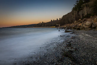 A serene coastal scene at dawn with soft mist rolling over calm waters beneath a pastel sky.