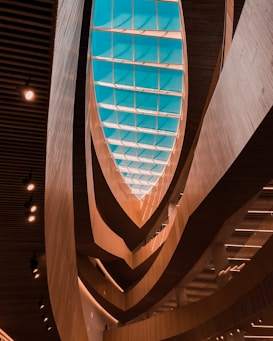 An architectural interior view featuring a large, curved wooden structure with layered patterns. A ceiling skylight in a geometric design allows natural light to filter through, casting a soft glow on the sleek wooden surfaces. Recessed lighting along the ceiling adds an additional source of illumination, enhancing the modern and sophisticated aesthetic.