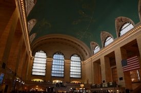 A large hall with a vaulted ceiling featuring intricate constellations and a blue-green color. Tall arched windows allow natural light to enter the space, and an American flag is prominently displayed. The interior architecture includes a combination of classical and luxurious stonework with a bustling atmosphere as numerous people move about the open area.