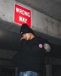 A person stands under a red 'WRONG WAY' sign on a concrete structure, wearing a black beanie and jacket. The image conveys a sense of urban exploration or rebellion.