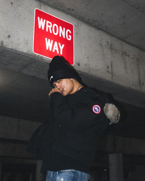A person stands under a red 'WRONG WAY' sign on a concrete structure, wearing a black beanie and jacket. The image conveys a sense of urban exploration or rebellion.