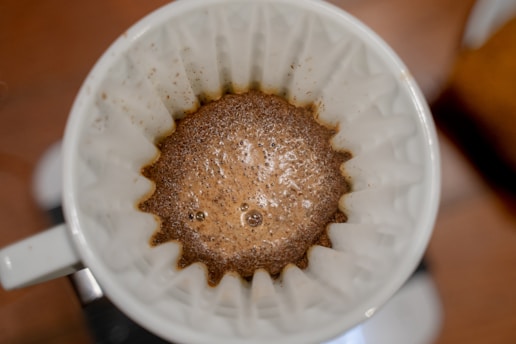 A close-up view of a coffee filter containing wet coffee grounds, likely in the process of brewing. The filter is pleated, and the coffee grounds appear to be soaked with water, forming a frothy layer on top. The surrounding area appears blurred.
