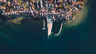 An aerial view of a coastal town with a densely packed arrangement of buildings near the shoreline. A marina extends into the water, with boats moored at docks. The scene is surrounded by calm, blue-green water, and the layout suggests a serene, organized community.