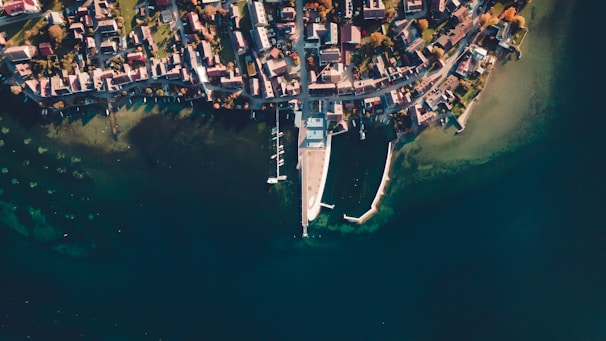 An aerial view of a coastal town with a densely packed arrangement of buildings near the shoreline. A marina extends into the water, with boats moored at docks. The scene is surrounded by calm, blue-green water, and the layout suggests a serene, organized community.