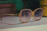 A pair of large, vintage-style sunglasses with a light brown frame and gradient lenses rests on a white surface. In the background, there are some blurred objects, including books and a yellow container.