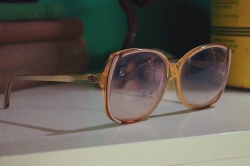 A pair of large, vintage-style sunglasses with a light brown frame and gradient lenses rests on a white surface. In the background, there are some blurred objects, including books and a yellow container.