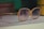 A pair of large, vintage-style sunglasses with a light brown frame and gradient lenses rests on a white surface. In the background, there are some blurred objects, including books and a yellow container.