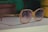 A pair of large, vintage-style sunglasses with a light brown frame and gradient lenses rests on a white surface. In the background, there are some blurred objects, including books and a yellow container.