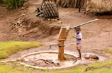 A young child operates a manual water pump, surrounded by a rural landscape. The area features bare, earthy ground with patches of grass, and a wooden structure in the background. The child is barefoot and wearing a light blue shirt and shorts.