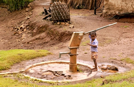 A young child operates a manual water pump, surrounded by a rural landscape. The area features bare, earthy ground with patches of grass, and a wooden structure in the background. The child is barefoot and wearing a light blue shirt and shorts.