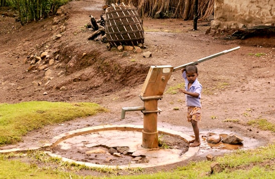 A young child operates a manual water pump, surrounded by a rural landscape. The area features bare, earthy ground with patches of grass, and a wooden structure in the background. The child is barefoot and wearing a light blue shirt and shorts.