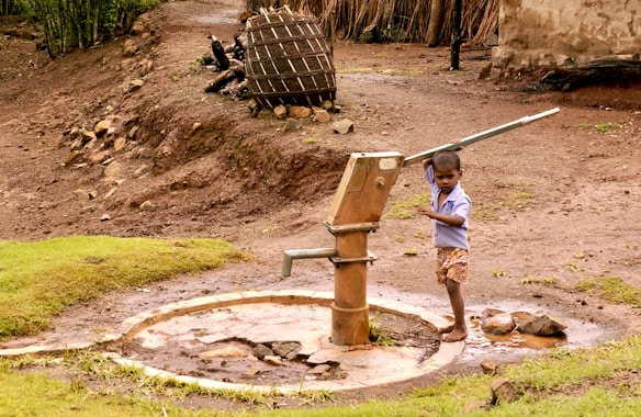 A young child operates a manual water pump, surrounded by a rural landscape. The area features bare, earthy ground with patches of grass, and a wooden structure in the background. The child is barefoot and wearing a light blue shirt and shorts.