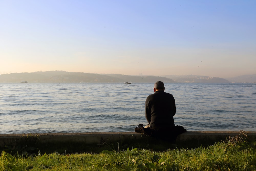 man in black jacket sitting on brown rock near body of water during daytime