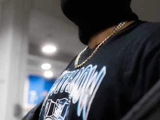 A person is wearing a black balaclava, a gold chain necklace, and a black shirt with white and blue graphics in a well-lit indoor setting. The background features several round light fixtures, a blue wall, and some blurred objects.