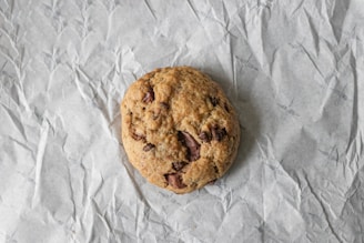 A single chocolate chip cookie rests on a crinkled piece of parchment paper. The cookie has a slightly golden-brown color with visible chunks of chocolate throughout.
