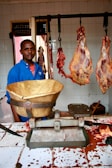 A man wearing a blue shirt stands in a butcher shop. There are large pieces of meat hanging from hooks behind him. A weighing scale is placed on the counter which is stained with blood. The walls are tiled and there is a window in the background.
