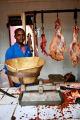A man wearing a blue shirt stands in a butcher shop. There are large pieces of meat hanging from hooks behind him. A weighing scale is placed on the counter which is stained with blood. The walls are tiled and there is a window in the background.