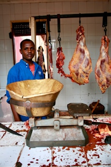 A man wearing a blue shirt stands in a butcher shop. There are large pieces of meat hanging from hooks behind him. A weighing scale is placed on the counter which is stained with blood. The walls are tiled and there is a window in the background.