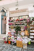 A flower shop is adorned with a vibrant display of various flowers in pots, baskets, and crates. The storefront is decorated with colorful floral arrangements and greenery. A large street lamp is mounted on the wall, and multiple signs are present with text. Several religious icons are visible in the window above the flowers.