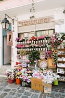 A flower shop is adorned with a vibrant display of various flowers in pots, baskets, and crates. The storefront is decorated with colorful floral arrangements and greenery. A large street lamp is mounted on the wall, and multiple signs are present with text. Several religious icons are visible in the window above the flowers.
